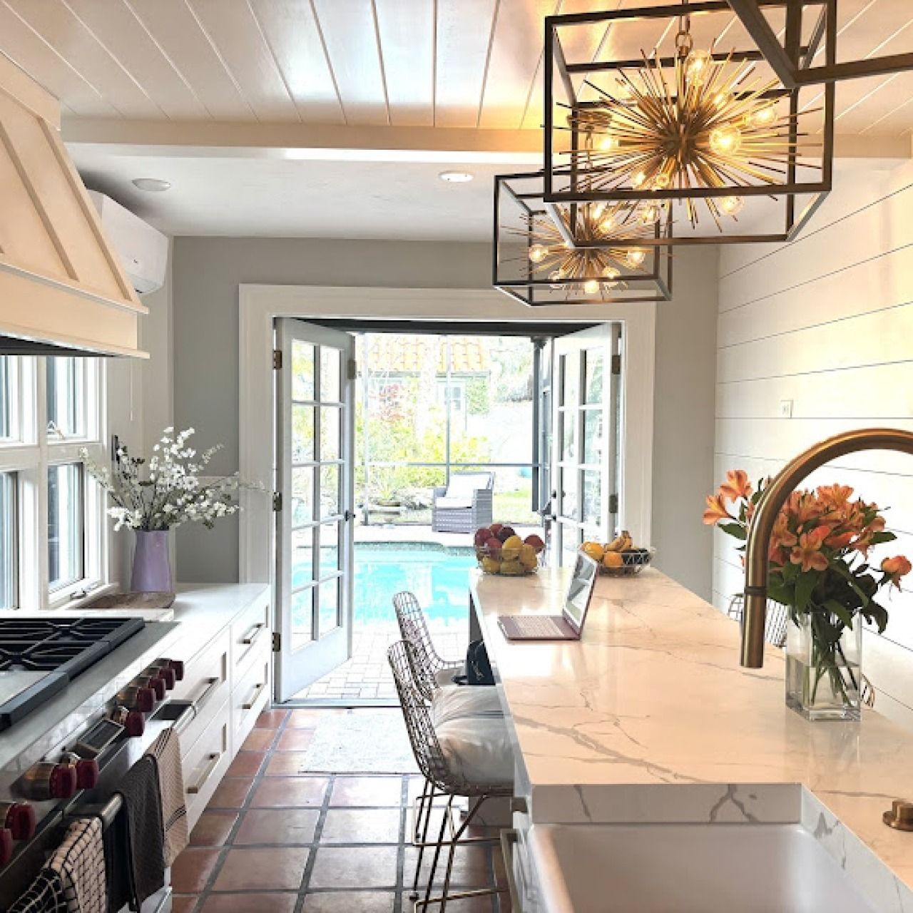 Bright kitchen with marble island, bar stools, terracotta floor, and open doors to a pool courtyard.