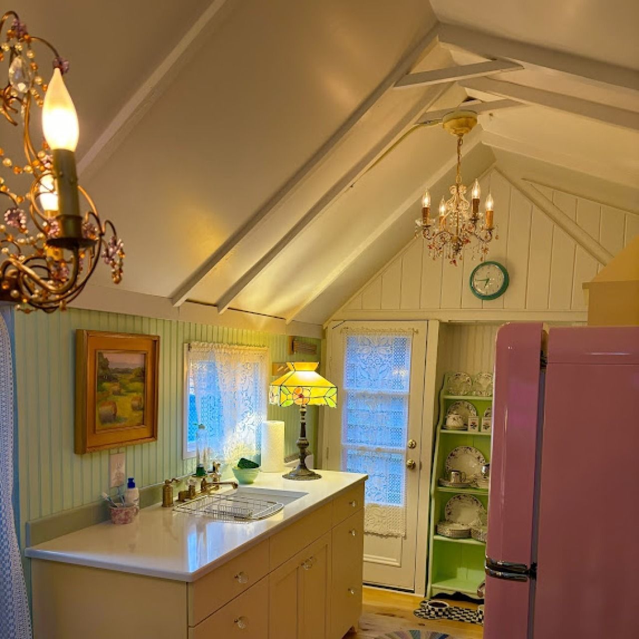 Cozy pastel kitchen with vaulted ceiling, chandelier, green cabinets, pink fridge, and white countertop
