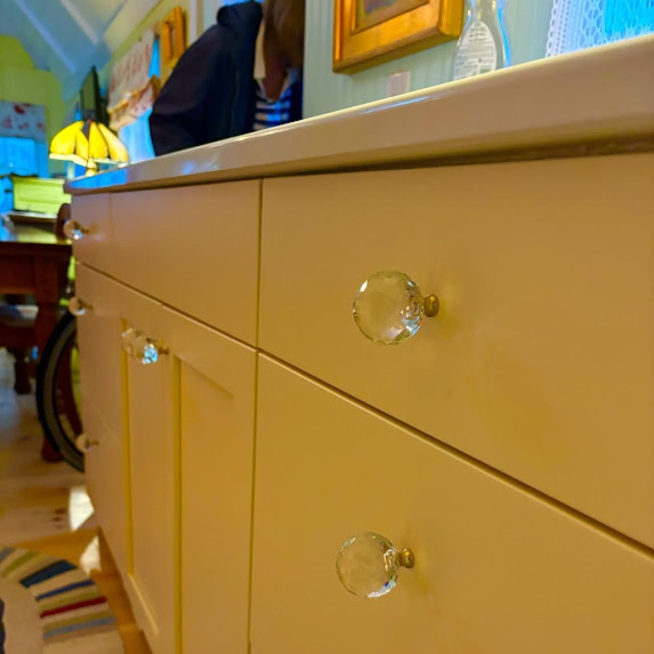 Cream cabinet with crystal knobs in a warmly lit room, with a chair and framed wall art in the background