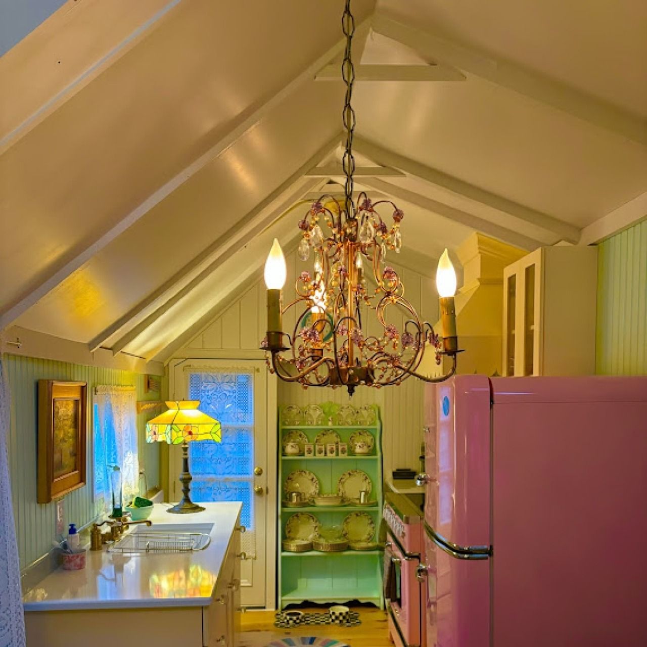 Cozy attic kitchen with pink refrigerator, white cabinets, chandelier, and yellow lamp lighting.