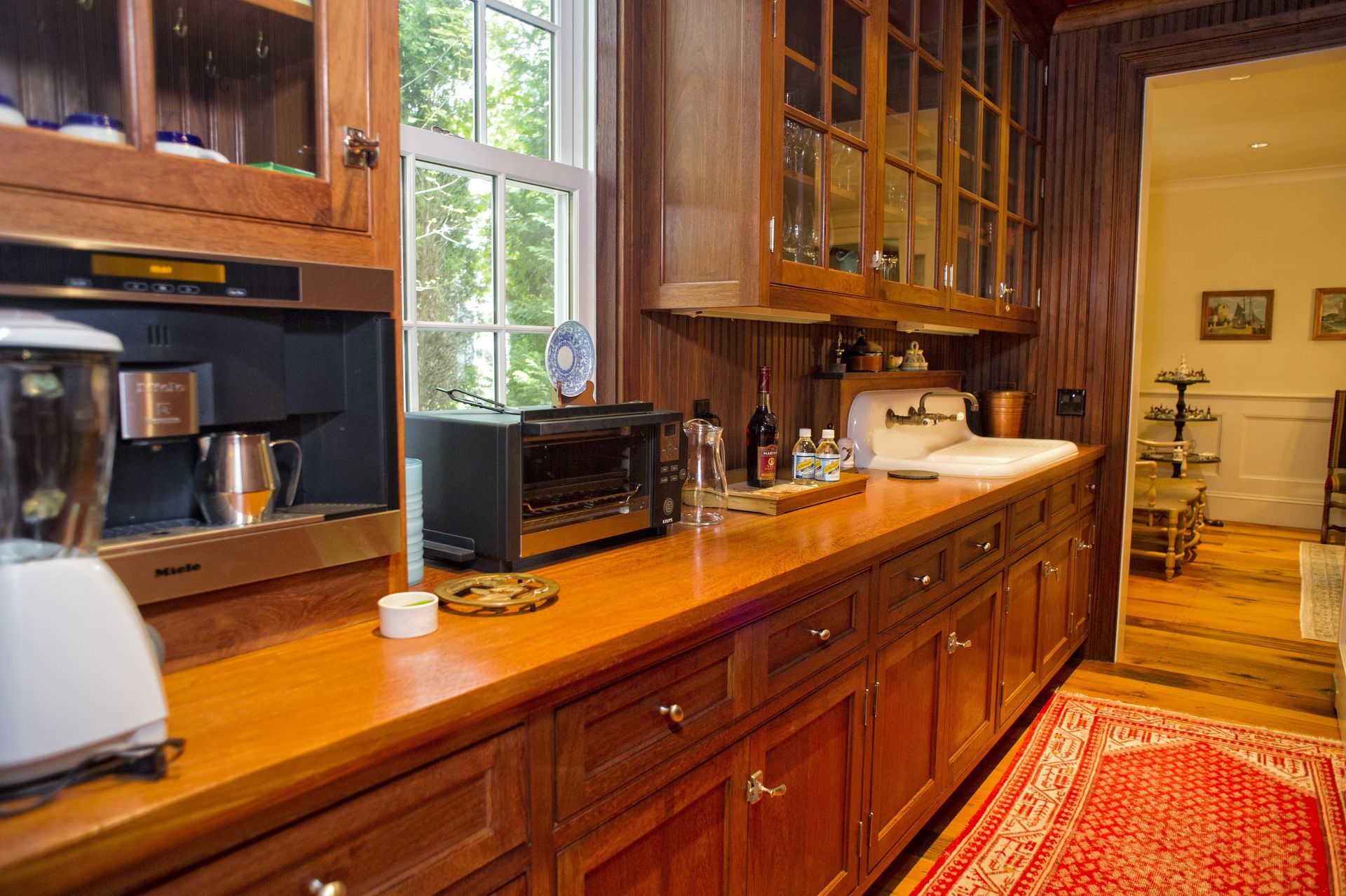 Wooden kitchen with a long counter, cabinets, window, toaster, microwave, and red rug leading to a hallway.
