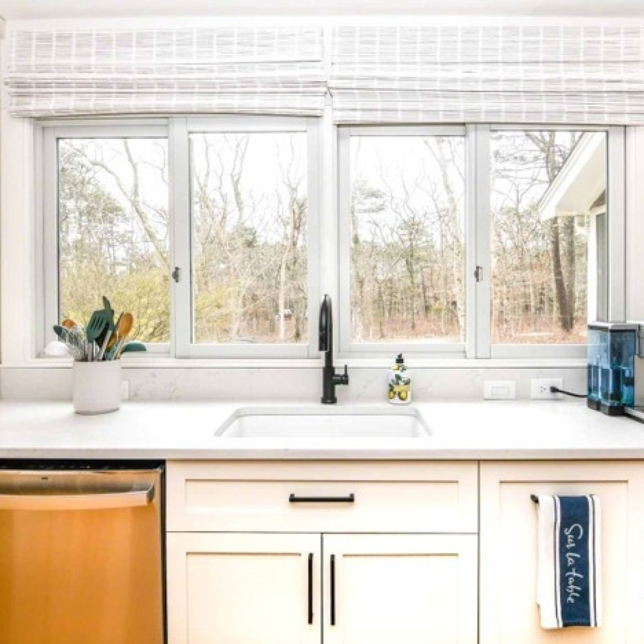 Bright white kitchen sink under large windows with a potted plant and orange dishwasher on the left