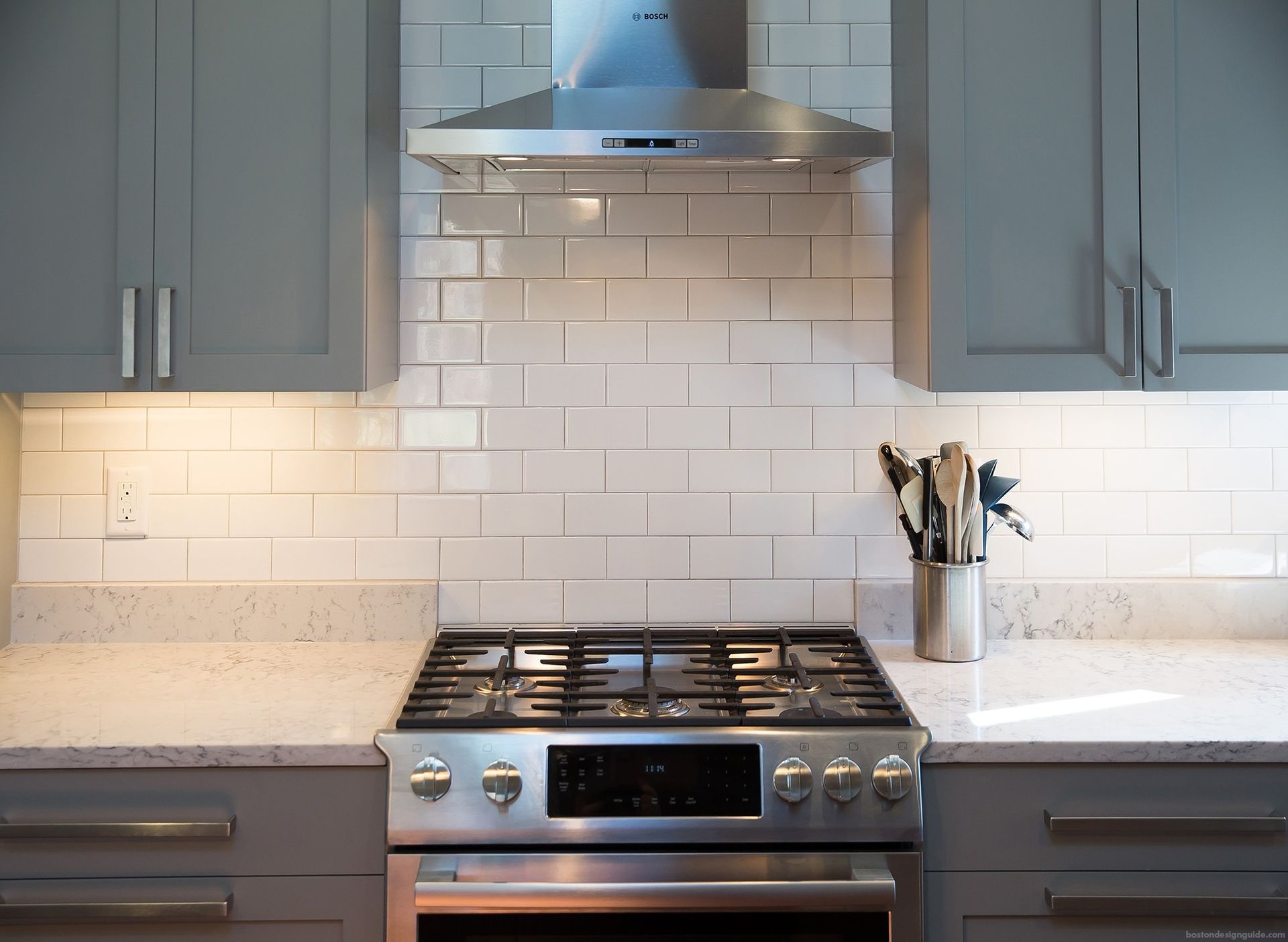 Modern kitchen with gray cabinets, white subway tile backsplash, stainless steel stove, and countertop utensils
