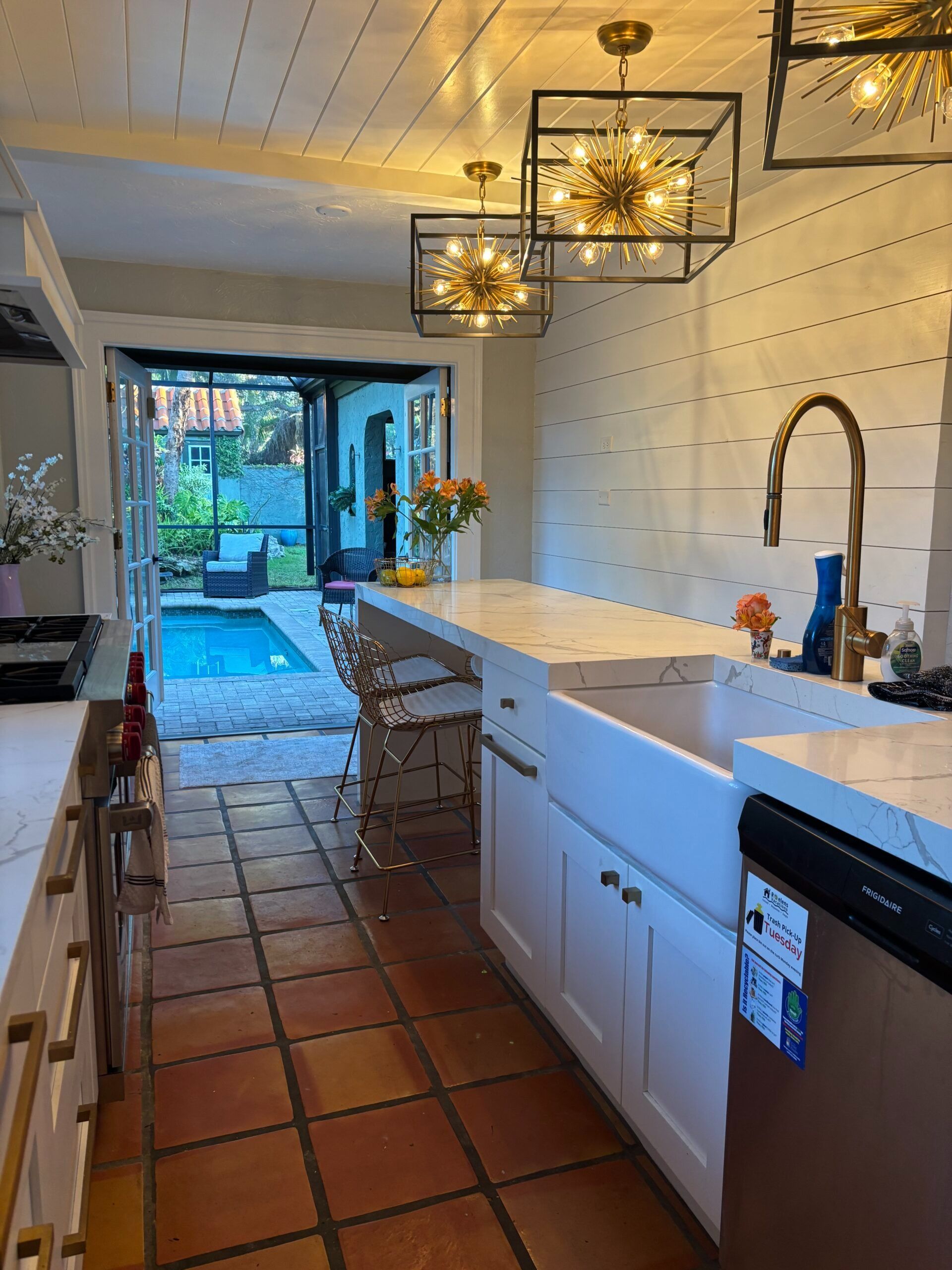 Long kitchen with terracotta floor, white cabinets, island, pendant lights, and open doors to a garden.