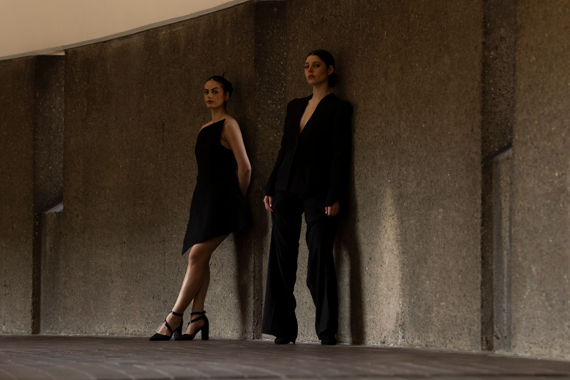 Two women in black outfits lean against a concrete wall. One in a dress, one in a suit.