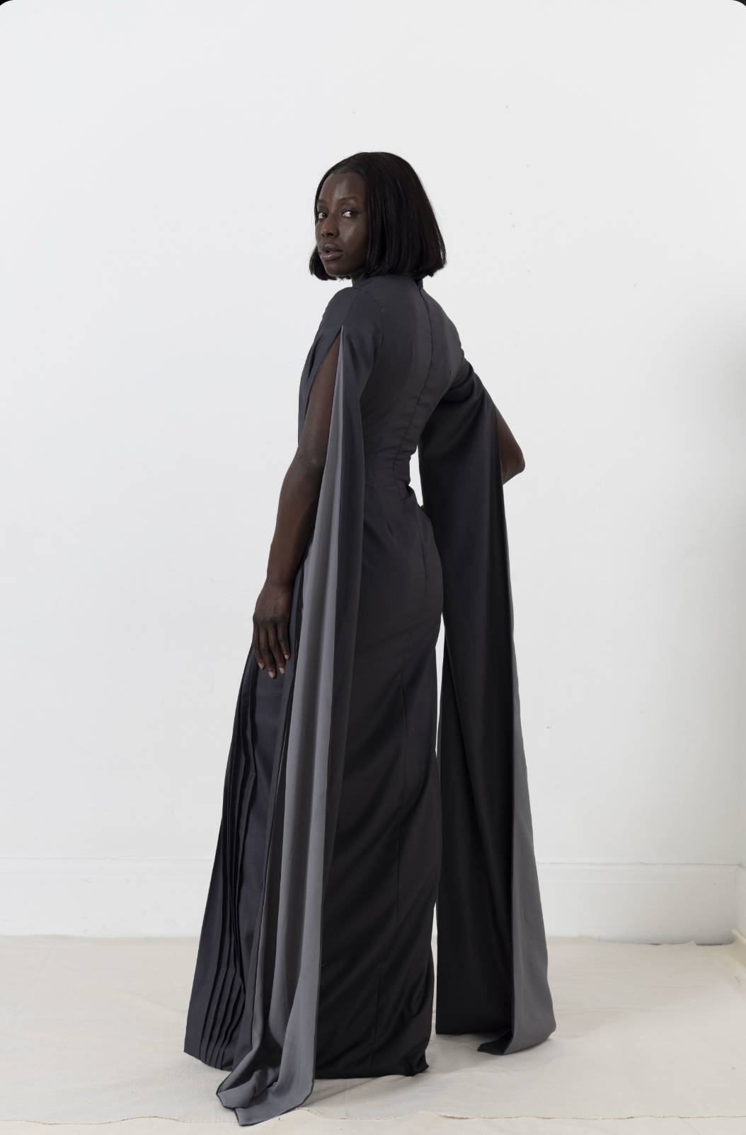 Woman in a dark gray gown with flowing sleeves, standing in a white room, looking over her shoulder.