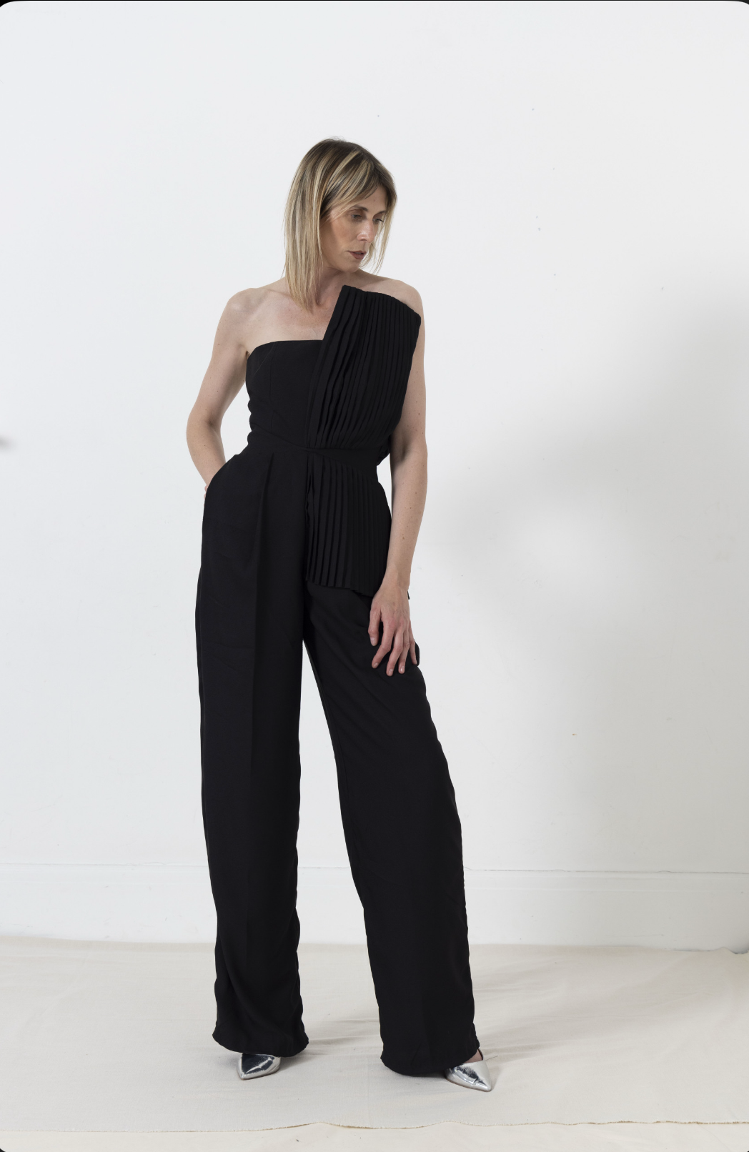 Woman in black strapless jumpsuit with pleated bodice, hands in pockets, standing against a white wall.