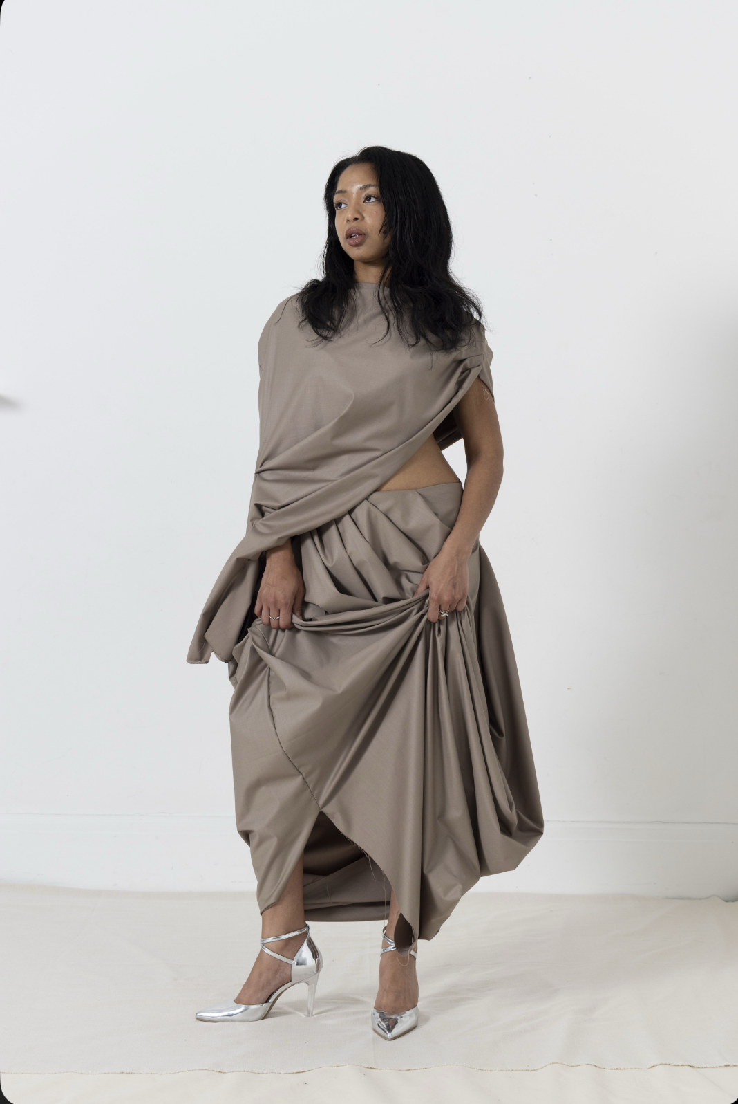 Woman in draped tan dress and silver heels, looking up in a white room.