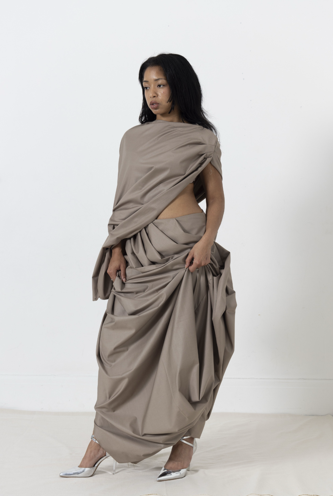 Woman in draped taupe dress and silver heels, standing against a white wall, looking left.