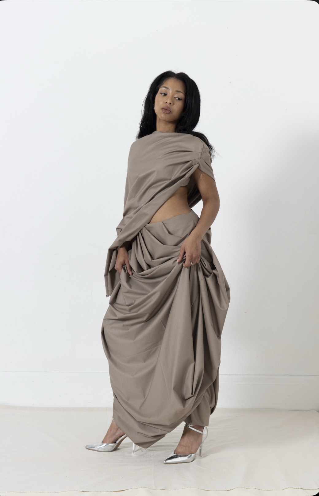 Woman in draped, taupe gown and silver heels posing against white backdrop.