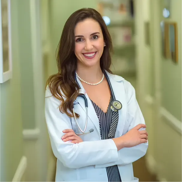 Woman in a white coat with a pearl necklace smiles, arms crossed, in a blurred office setting.