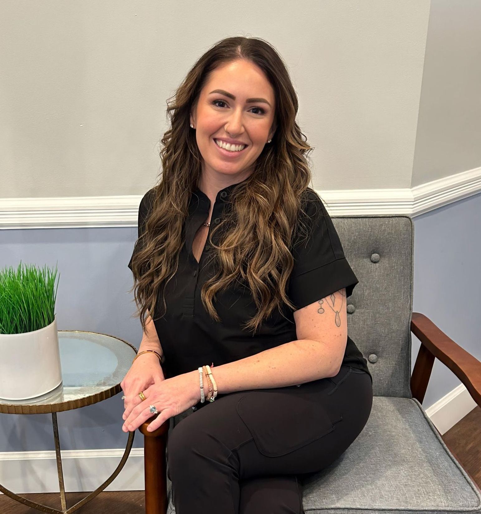 Woman smiling, seated, wearing black scrubs. Beside her, a table with a plant. Pale blue wall.