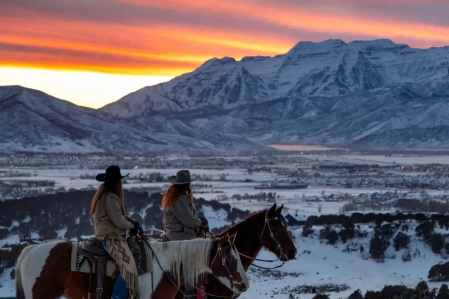Two people riding horses in the snow with mountains in the background