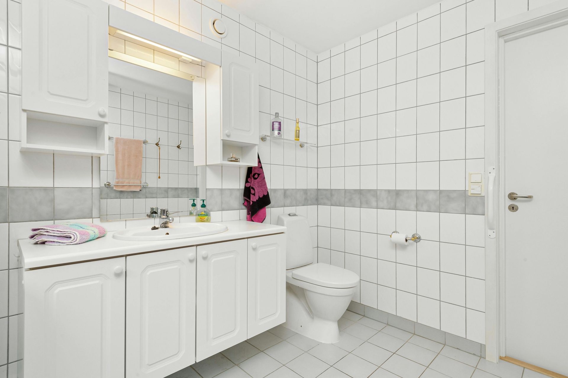 A clean, bright bathroom with white tiled walls, a white vanity cabinet, a sink, and a toilet.