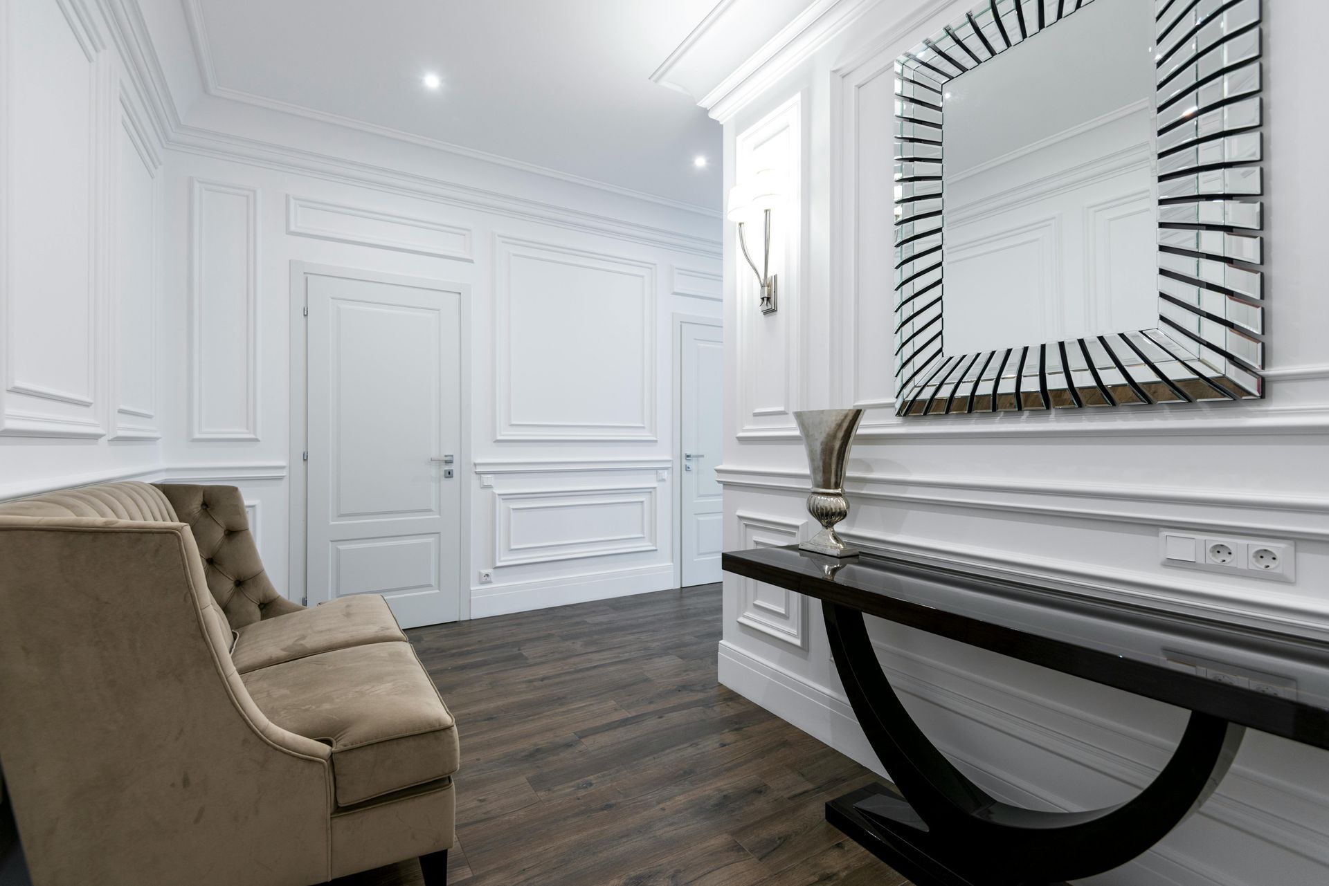A white hallway with paneled walls, a beige tufted sofa, a black console table with a silver vase, and a sunburst mirror.