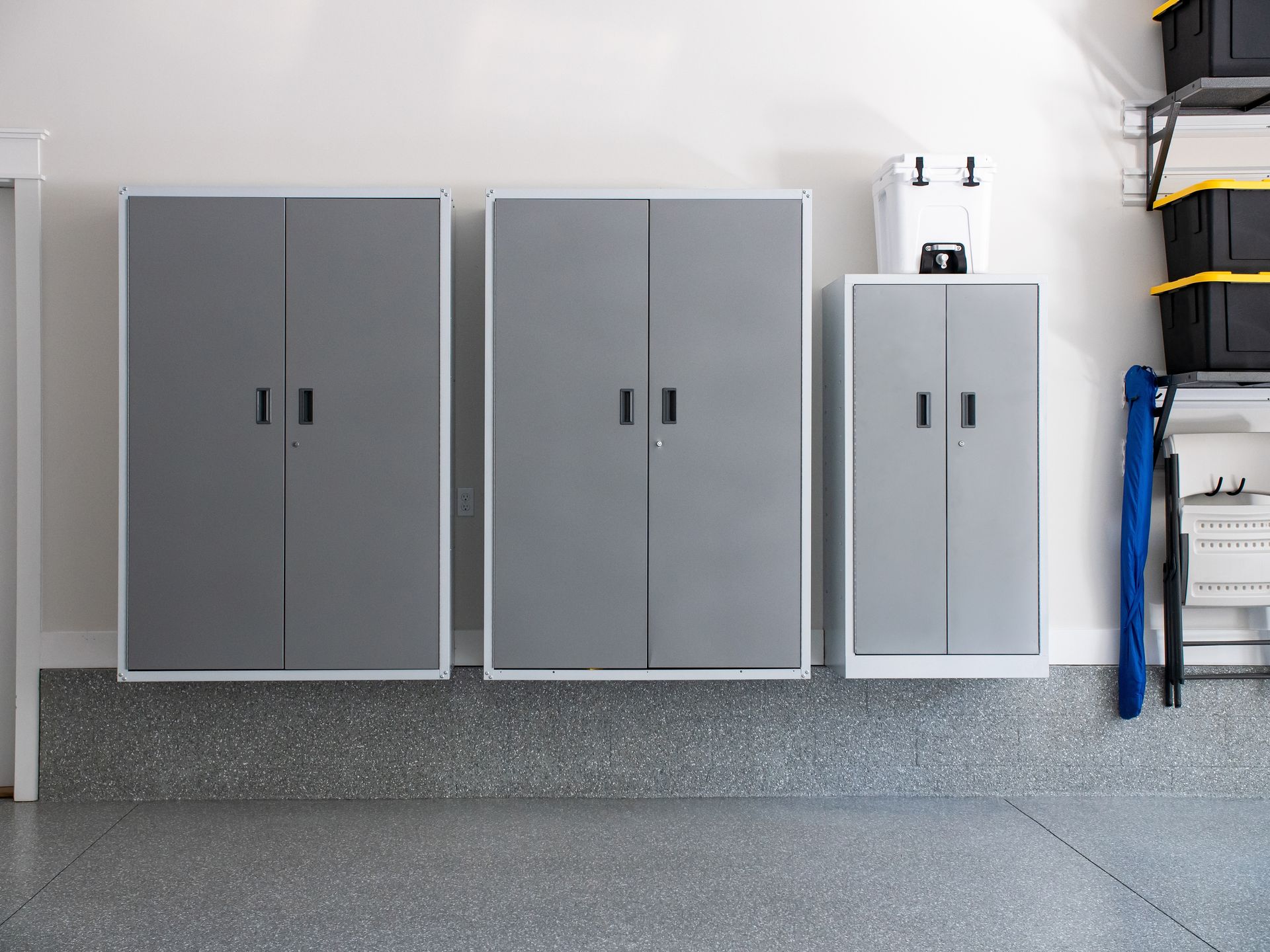 Three gray metal cabinets mounted on a garage wall above a speckled epoxy floor, with storage bins to the right.