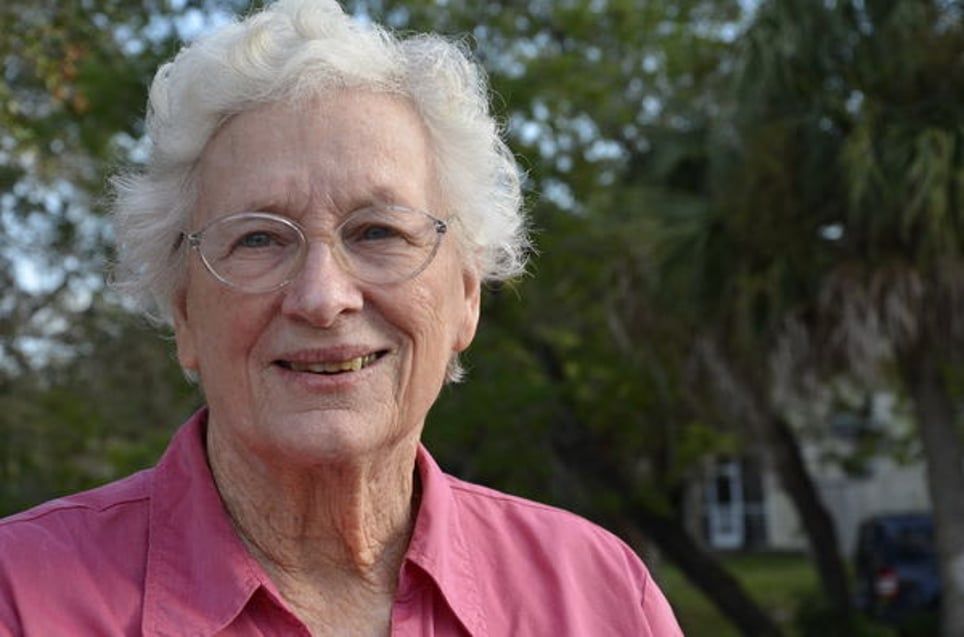 An elderly woman wearing glasses and a pink shirt is smiling for the camera.