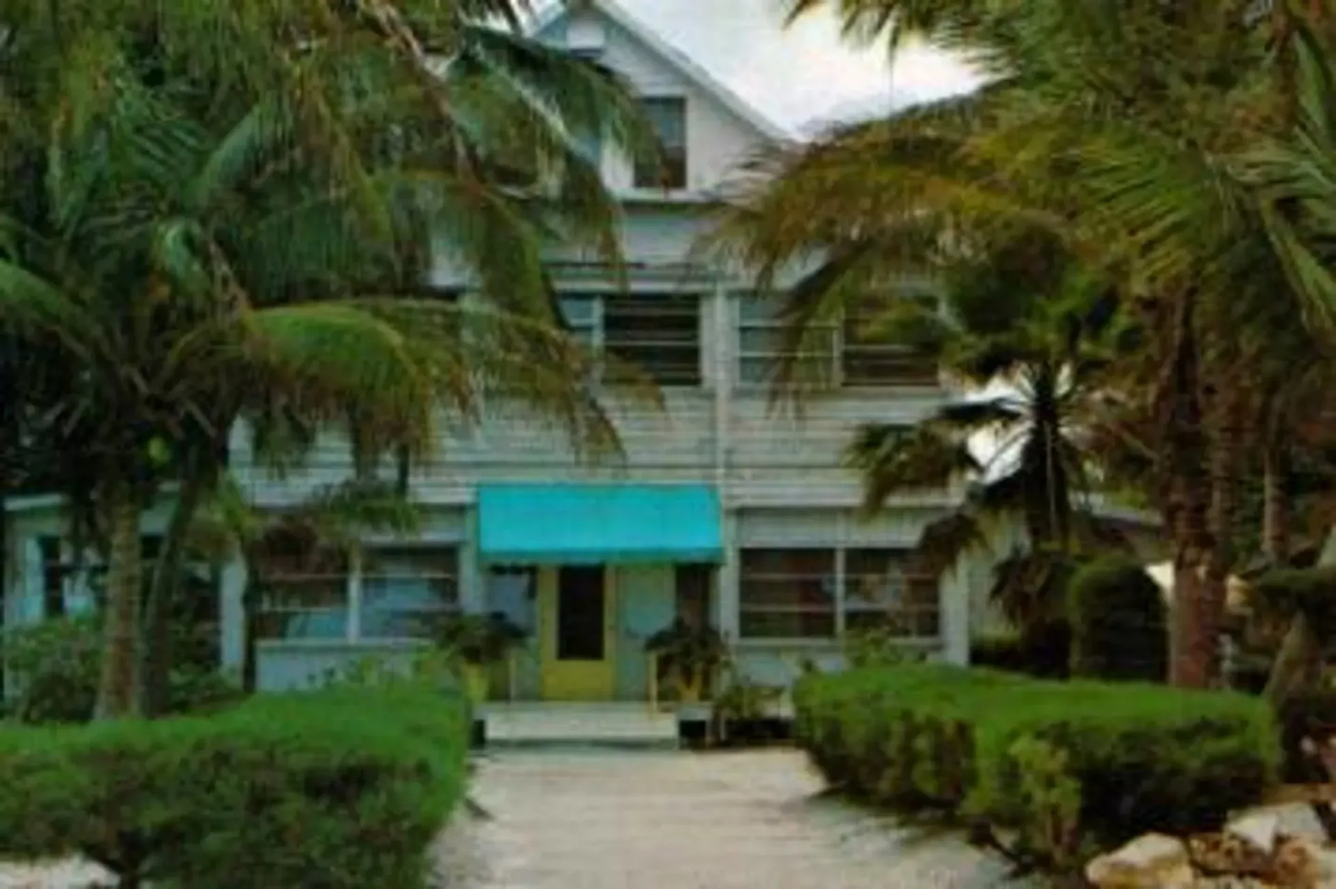 Tropical beachfront house with a turquoise awning, palm trees, and a sandy path leading to the entrance