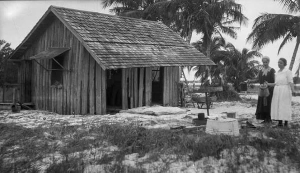 A black and white photo of two women standing in front of a small wooden house.
