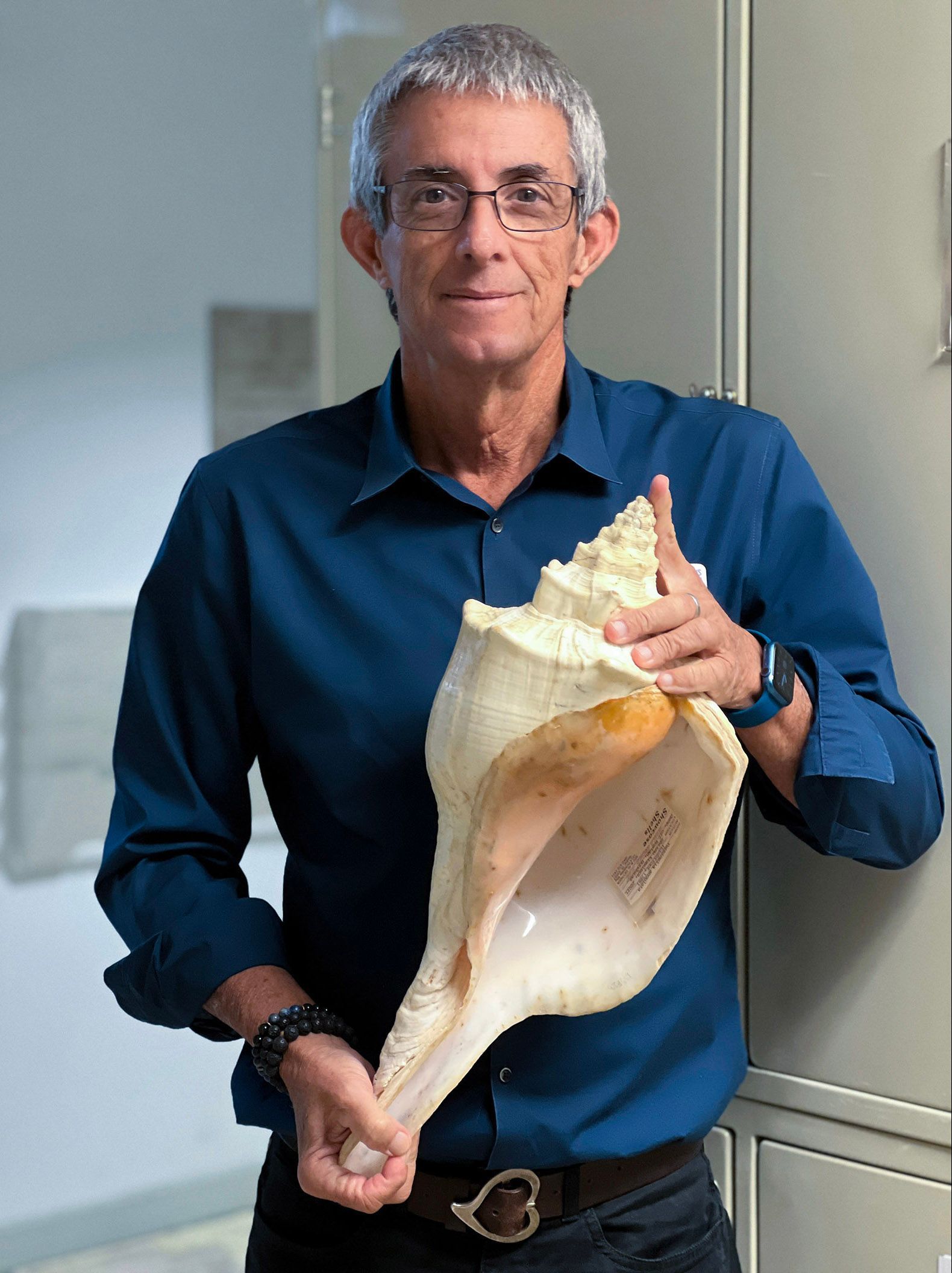 Man holding a large conch shell indoors, wearing a blue shirt and smiling.
