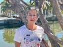 Woman with short gray hair, white t-shirt, standing by a body of water, smiling in the sun.