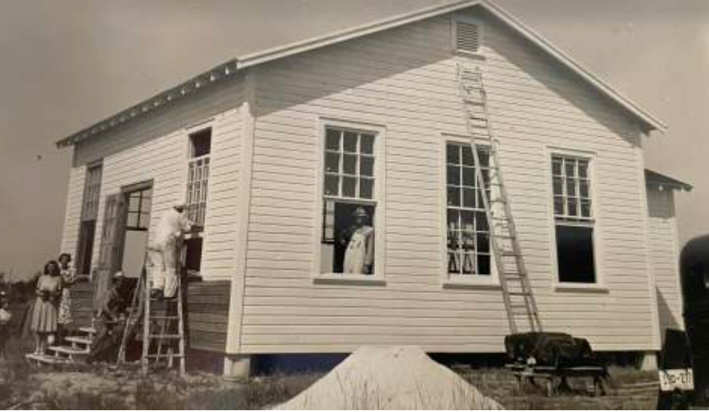 Black and white photo of a white clapboard building under construction. People stand in the windows and by a ladder.