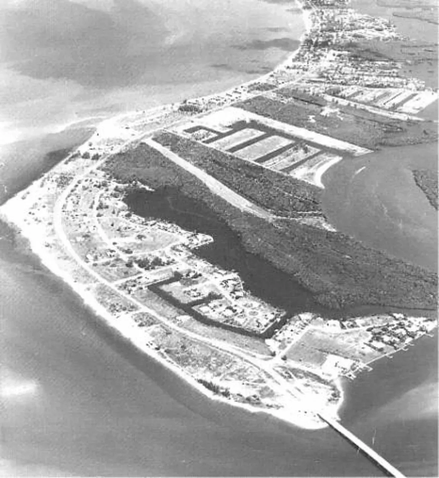 Aerial black-and-white view of a coastal island with buildings, shoreline, and a bridge or pier.