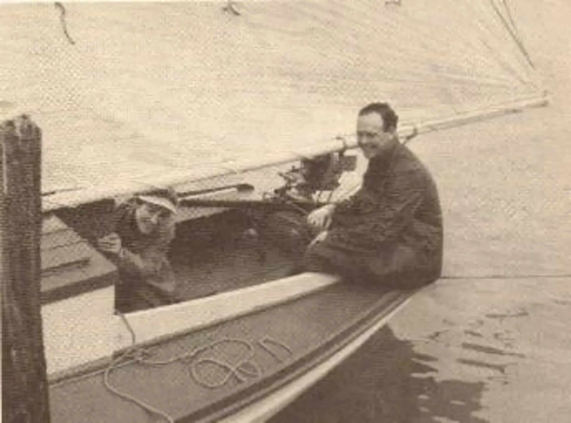 Two people in a small boat on calm water, one seated near the stern and one at the bow under a sail.