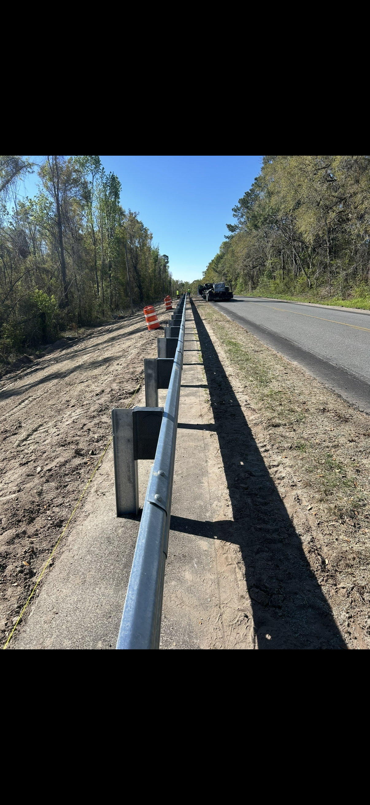 Roadside guardrail along a gravel shoulder beside trees and a clear blue sky.