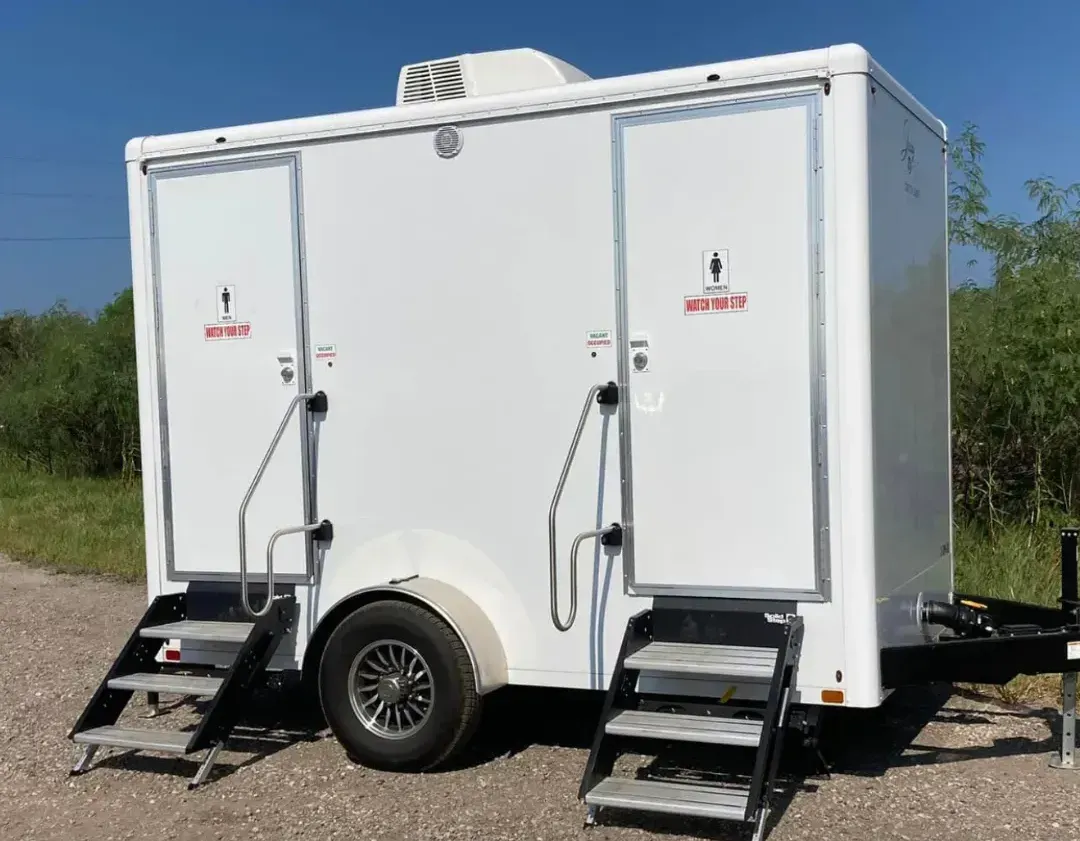 A white trailer with two toilets on it is parked on the side of a dirt road.