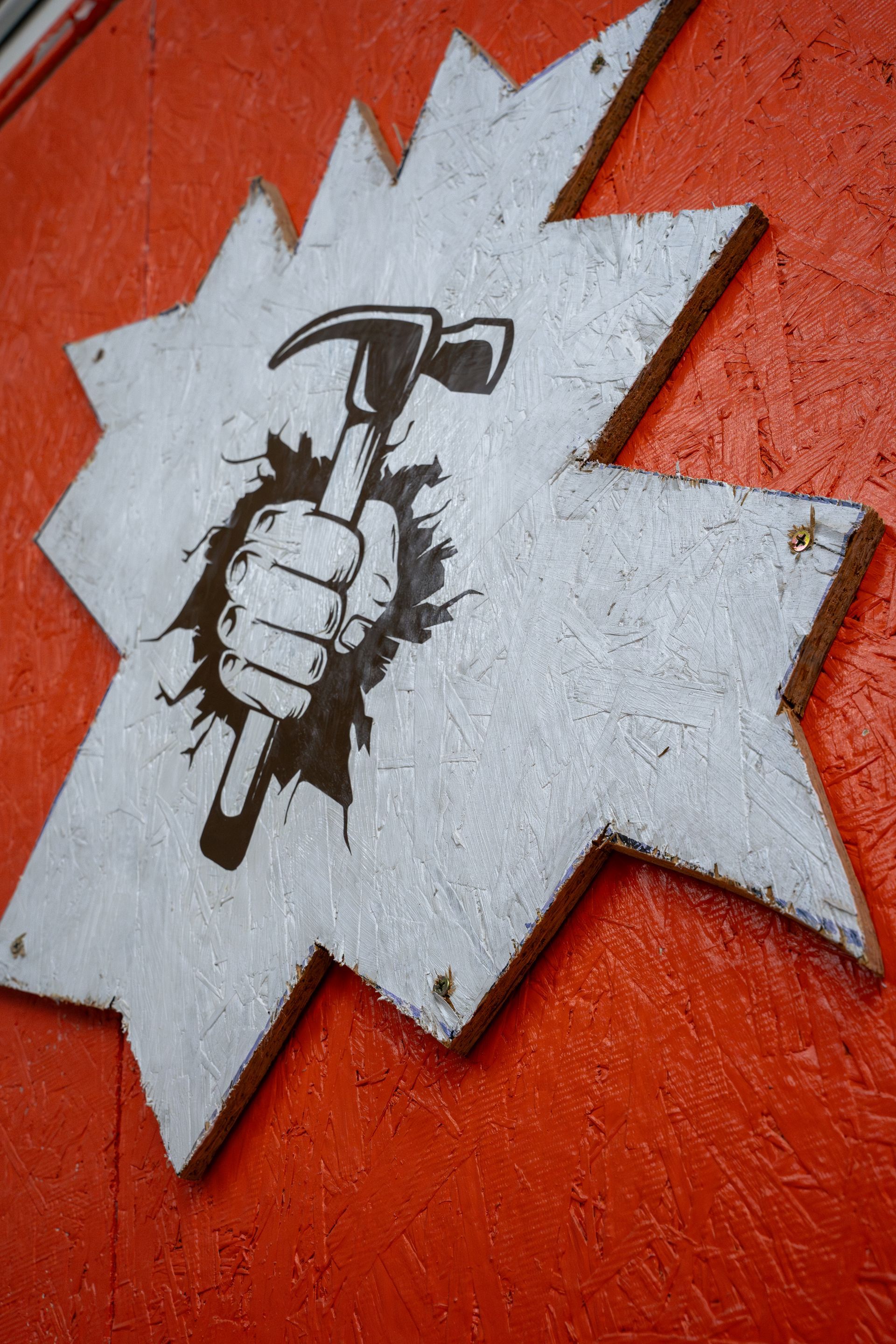 A white, star-shaped sign with a fist holding a hammer breaking through it; set against a red background.