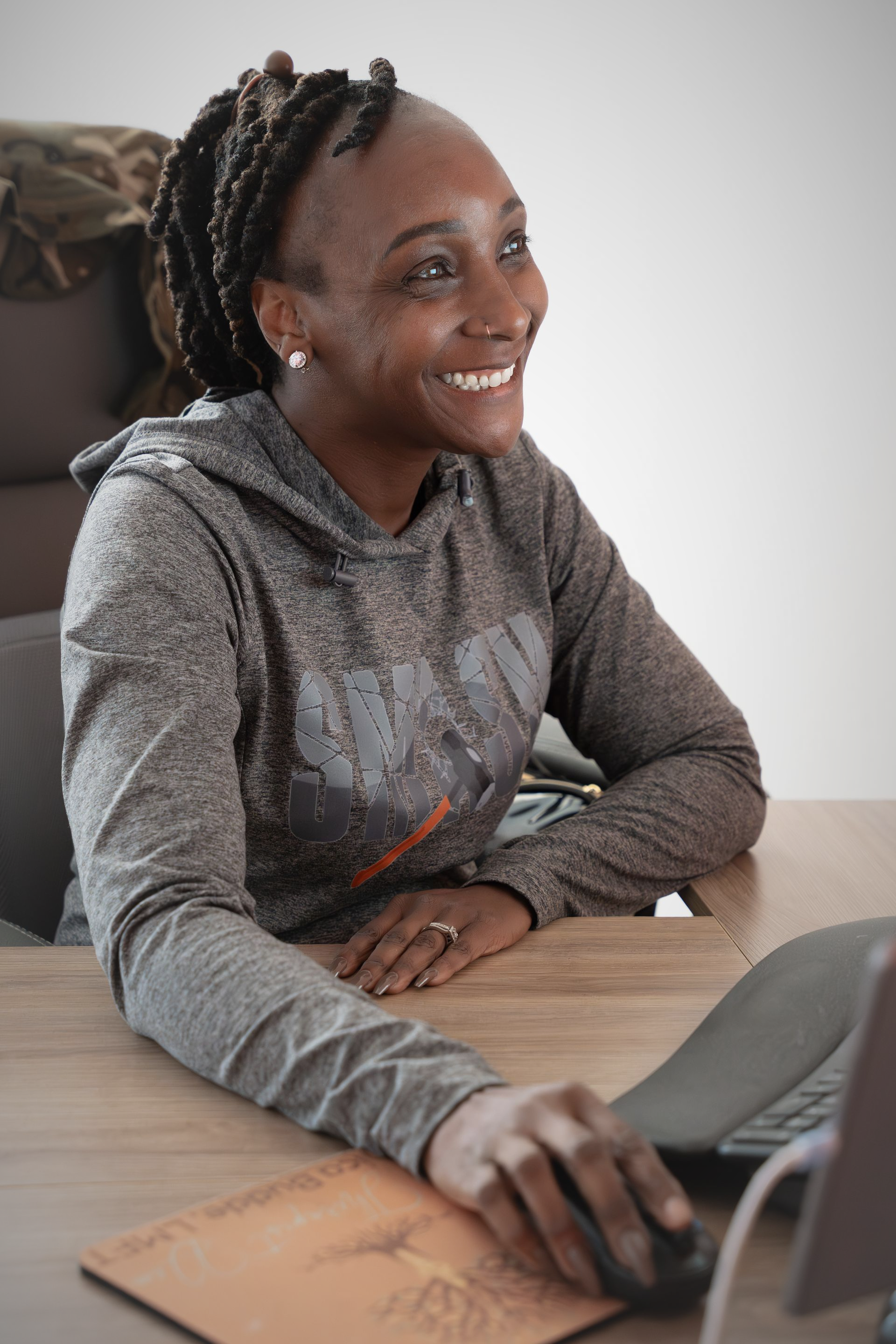 Woman smiling, working at desk with laptop and mouse, wearing grey hoodie.