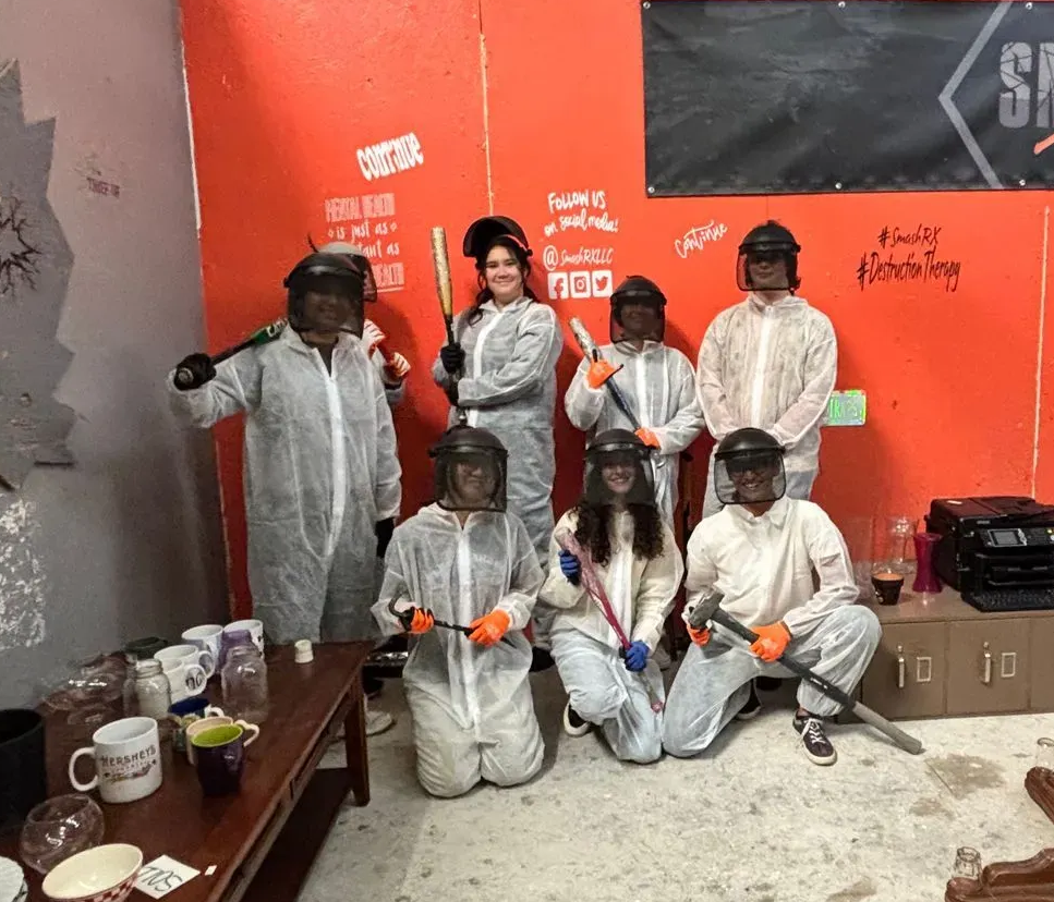 Group of people in protective gear pose in a red-walled smash room.