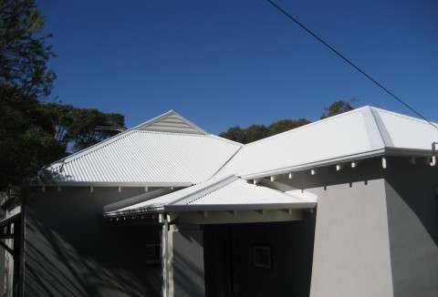 White-roofed house exterior under a clear blue sky, with trees and a driveway shadow in front.