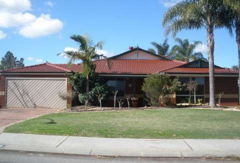 Single-story suburban house with a red tile roof, driveway, lawn, and palm trees under a blue sky