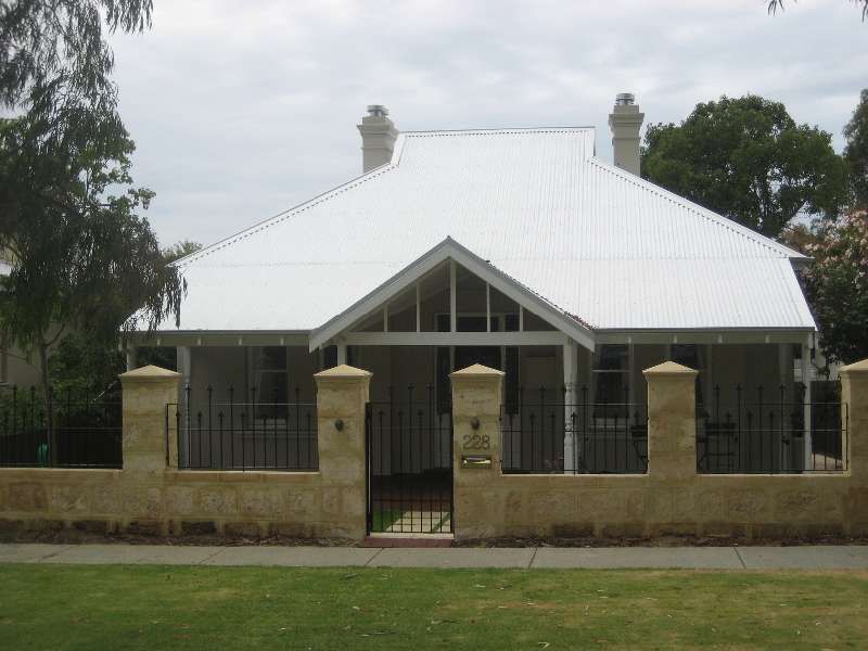 Small white house with a steep roof behind a stone fence and gate, framed by trees