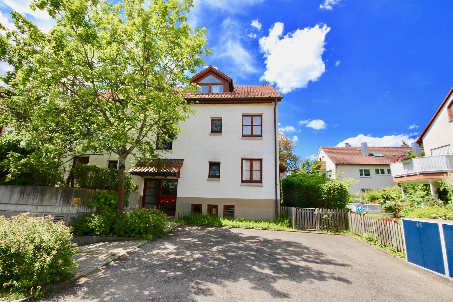 Sonniges Wohnhaus mit weißer Fassade, Baum davor und gepflasterter Auffahrt unter blauem Himmel