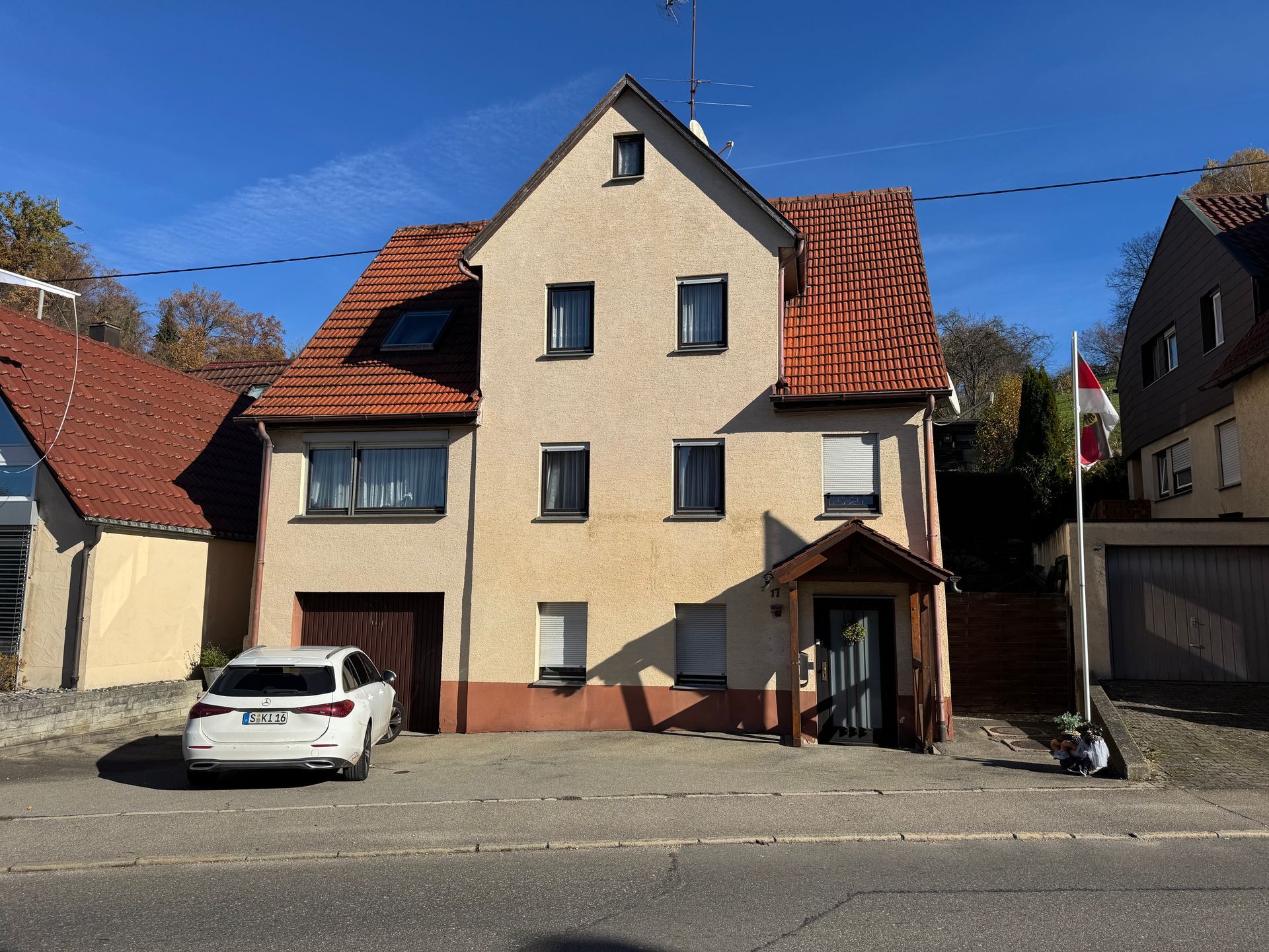 Ein cremefarbenes Haus mit rotem Ziegeldach und einem weißen Auto, das davor in einer sonnigen Straße parkt.