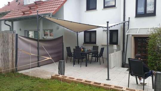 Terrasse mit Esstisch, Stühlen und beigefarbenem Sonnensegel neben einem Haus und einem umzäunten Garten