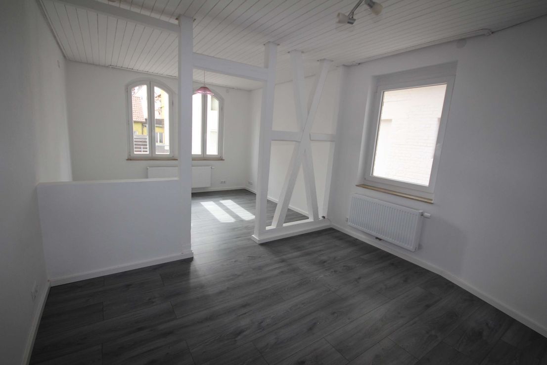 Ein leerer, weißer Raum mit dunkelgrauem Boden, großen Fenstern und einem Heizkörper an der Wand.
