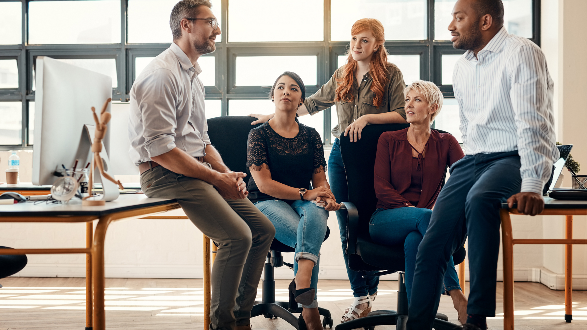 Four coworkers in a bright, modern office gathered for a discussion, with some seated and some standing.