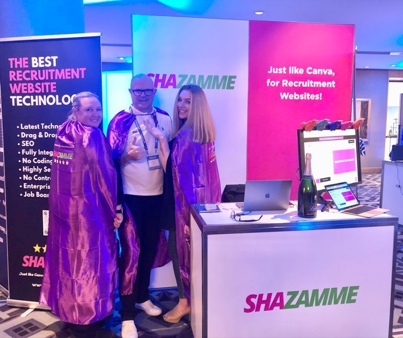Three people in purple capes stand behind a branded booth for Shazamme, a recruitment website platform.