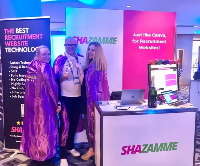 Three people in purple capes stand at a SHAZAMME branded booth with laptops and a bottle of champagne at an event.