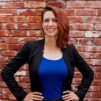 A person with auburn hair in a black blazer and blue shirt posing against a red brick wall.
