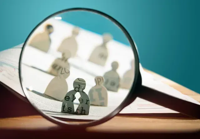 A magnifying glass focuses on one paper cutout person among many, highlighting the chosen individual.