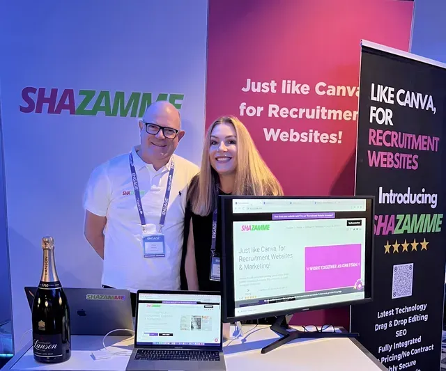 Two people stand at a SHAZAMME branded booth with a computer display and a bottle of champagne at a recruitment event.