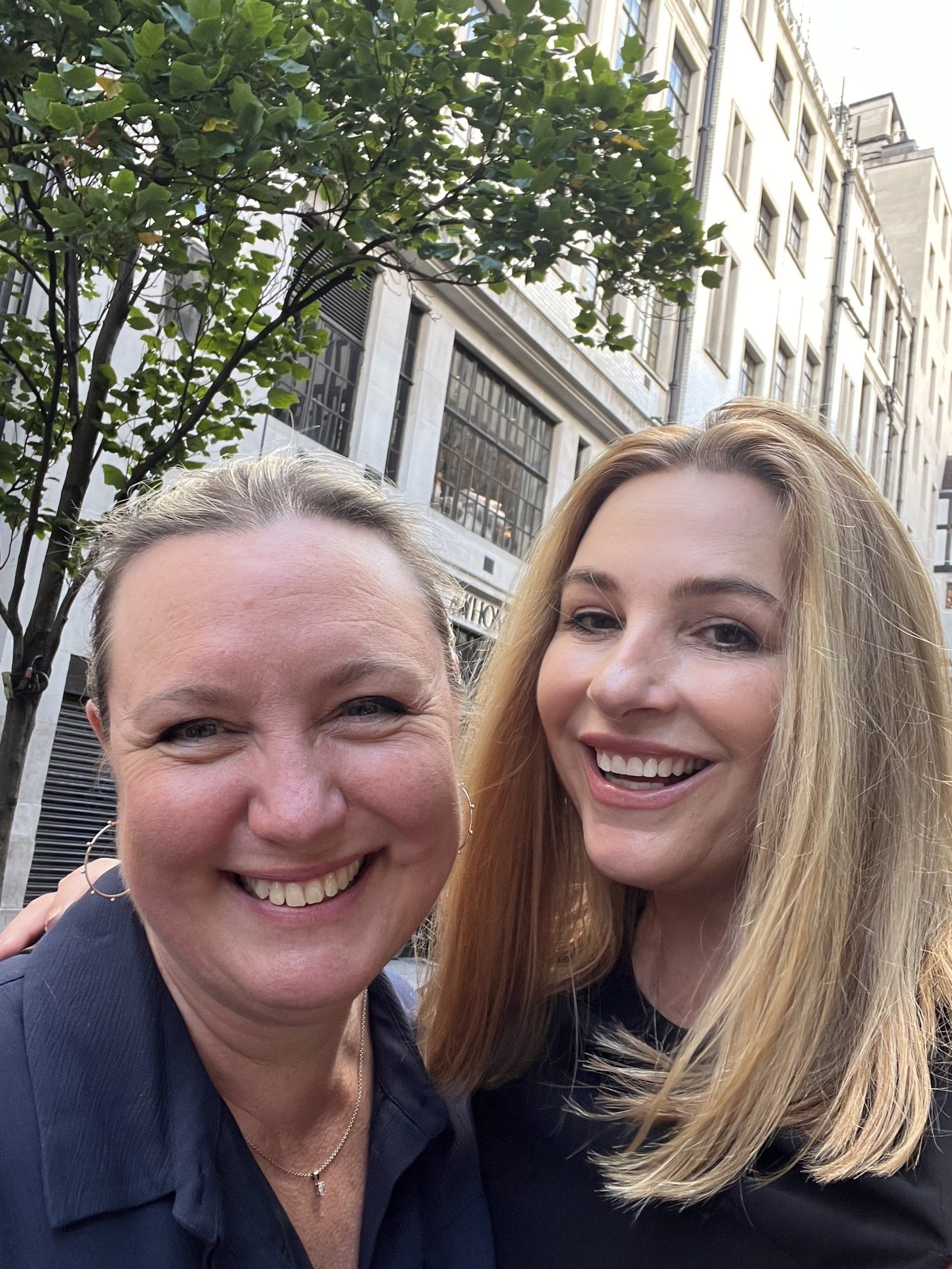 Two smiling people stand outdoors in front of a tree and a light-colored building.
