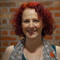 A smiling person with curly, bright red hair, wearing a patterned grey scarf, against a blurred brick wall background.