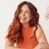 A person with wavy, reddish-brown hair smiles warmly while wearing an orange sleeveless top against a white background.