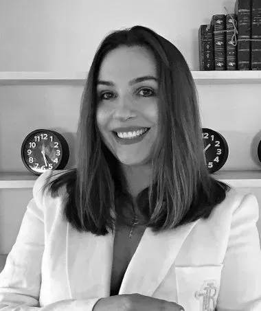 A person with shoulder-length hair smiles in a white blazer, standing in front of shelves holding books and clocks.